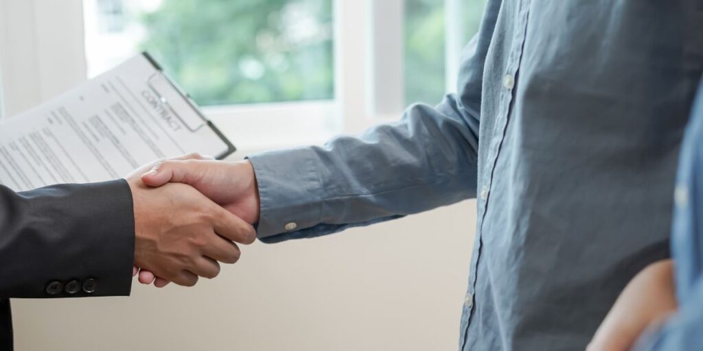 Close-up of two people shaking hands in an office, with one person holding a clipboard titled "CONTRACT" in the background.