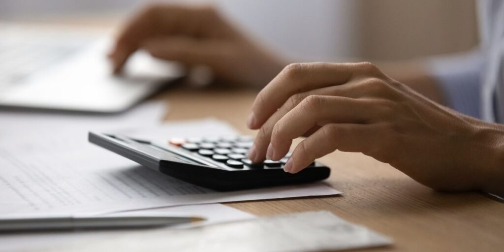 Close-up of hands using a desk calculator next to financial papers and a laptop in an office setting.